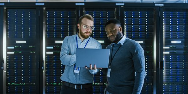 Bearded IT Technician in Glasses with a Laptop Computer and Black Male Engineer Colleague are Talking in Data Center while Working Next to Server Racks. Running Diagnostics or Doing Maintenance Work.